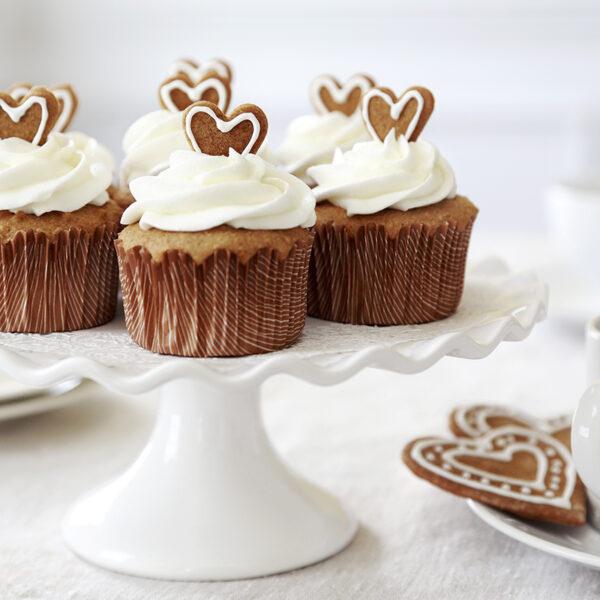 Gingerbread cupcakes with white piped icing and gingerbread hearts on top, displayed on a white cake stand, and a cup of coffee on the side in a white table setting