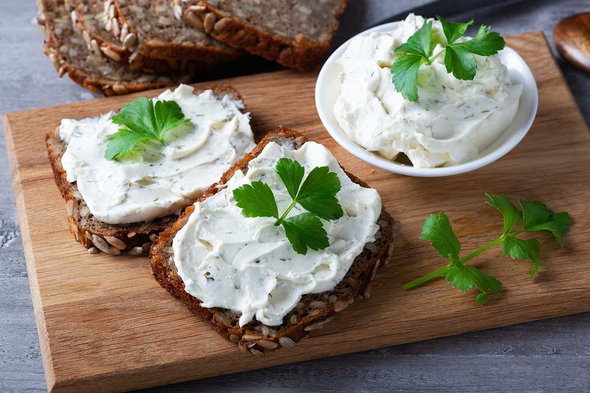 Herbed Ricotta Spread - sunflower-seed-studded bread, sliced and spread with white cheese mixed with green herbs. Parsley garnish. Wooden cutting board on grey cement surface.