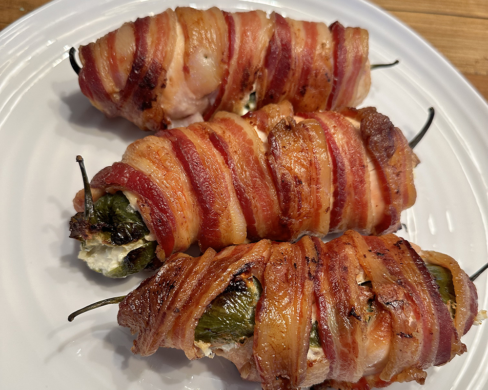 "Armadillo Eggs," cooked, bacon wrapped around jalapeno peppers stuffed with cream cheese. Three on a white plate.