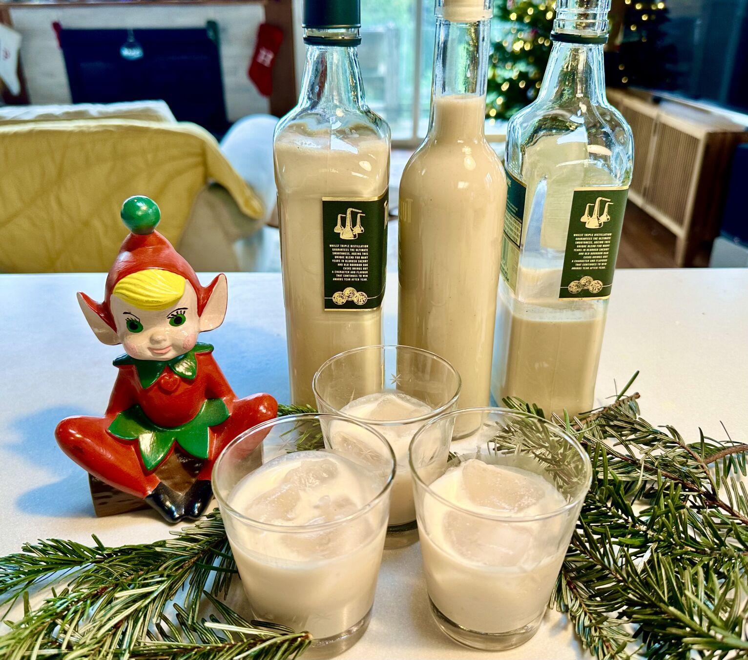 Leprechaun figurine sitting next to three bottles of tan-colored homemade Irish Cream, and three cocktail glasses with ice and Irish Cream. Sitting on white table with pine branches for decoration. Christmas decorations in background.