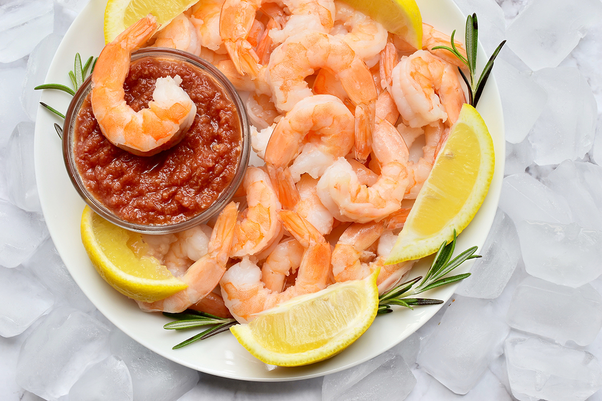 Shrimp Cocktail - Platter of cooked shrimp with lemon wedges, rosemary sprigs and small round bowl of cocktail sauce.