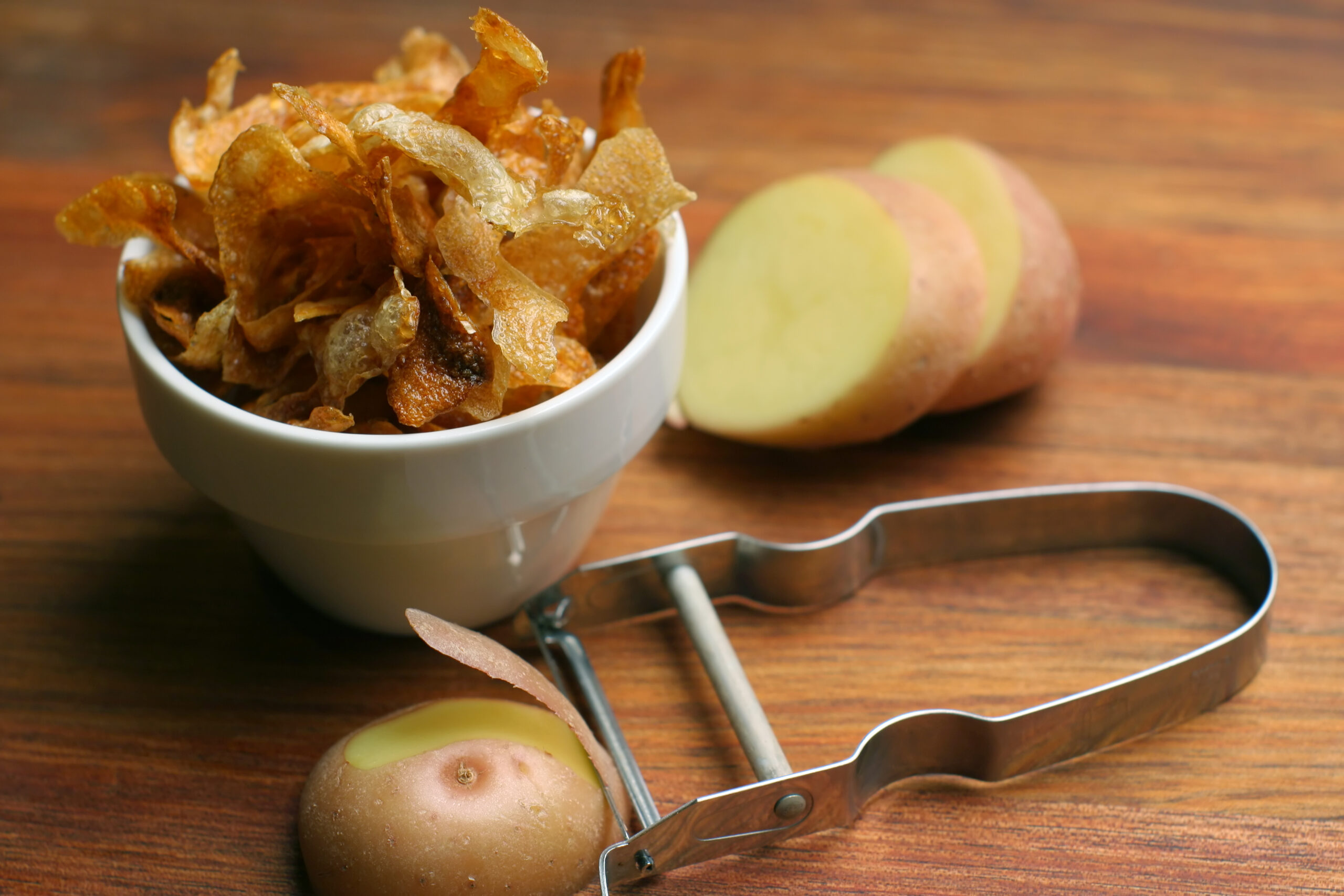 Crispy Oven-Roasted Potato Peels served in a white ceramic bowl, on wooden chopping block with peeler and potato.