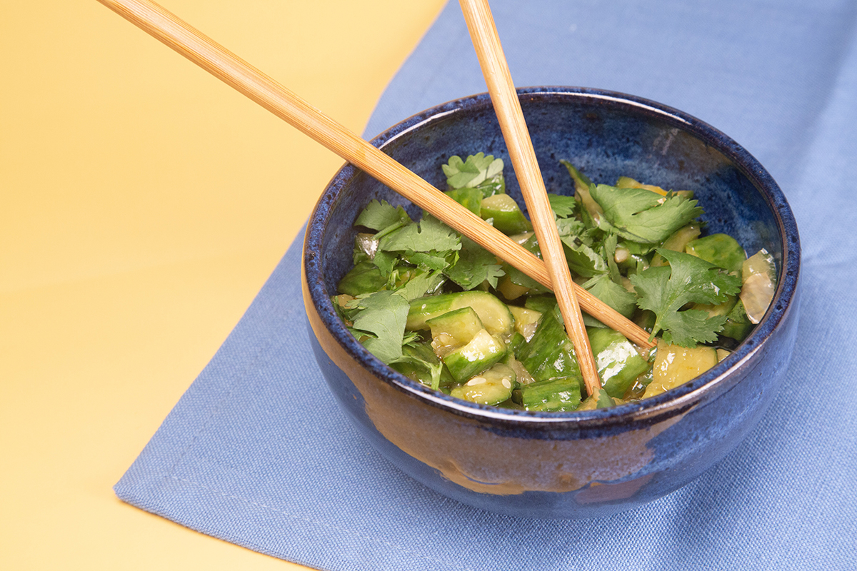 Spicy Smashed Cucumber Salad - Cucumber chunks mixed with vinagrette dressing and cilantro leaves. Served in a blue ceramic bowl with chopsticks, on blue and yellow table.