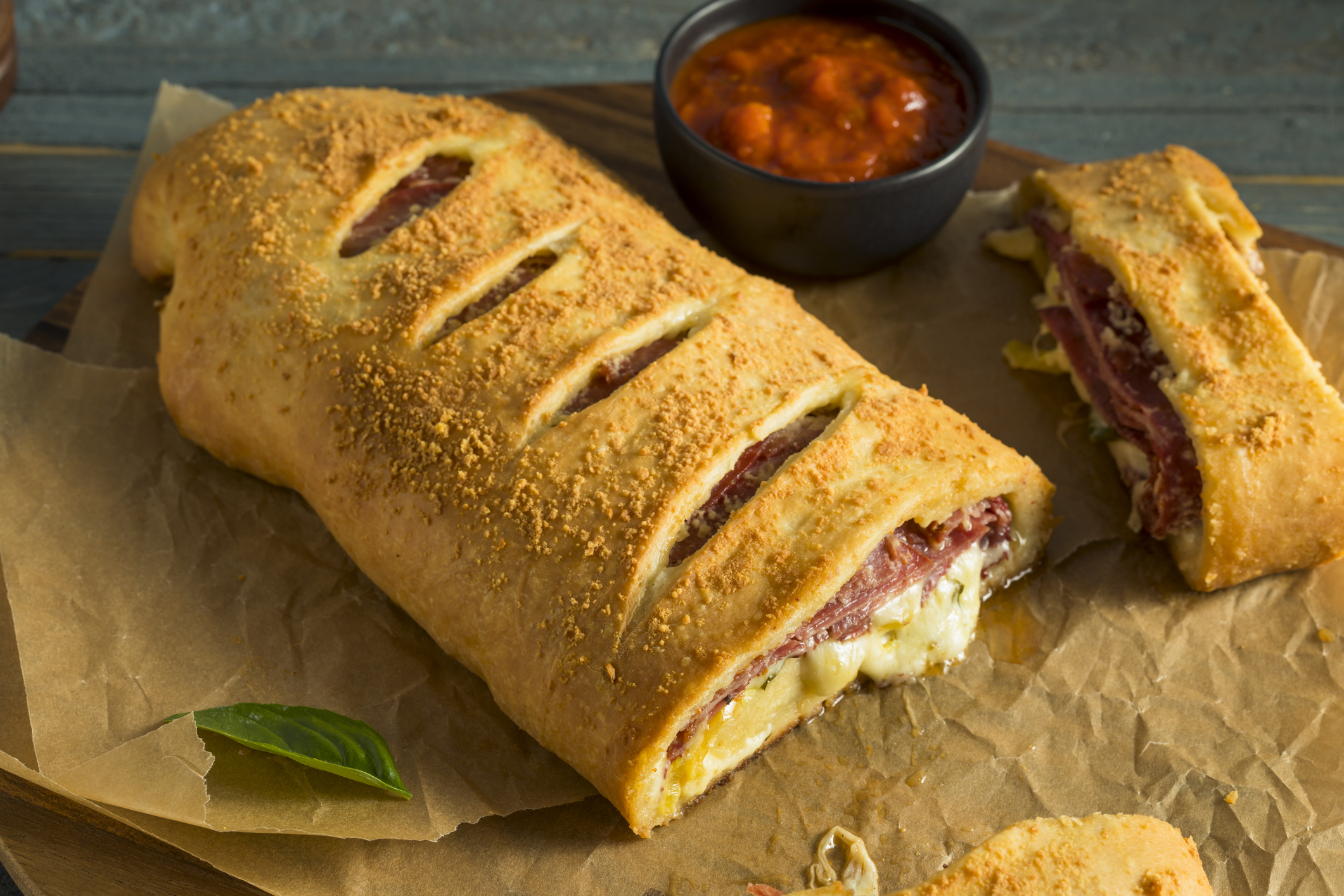 "Garbage Bread" - rolled stromboli-like bread with meat and cheese filling. sitting on brown paper bag with tomato dipping sauce in small black bowl.