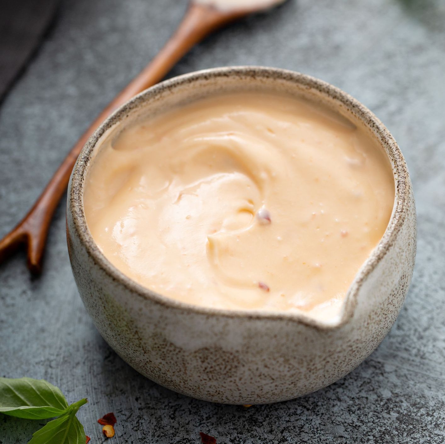 Homemade creamy dressing/sauce. Light orange-pink in a gray ceramic sauce bowl, accompanied by wooden spoon on a dark gray surface with herbs and chili flakes.
