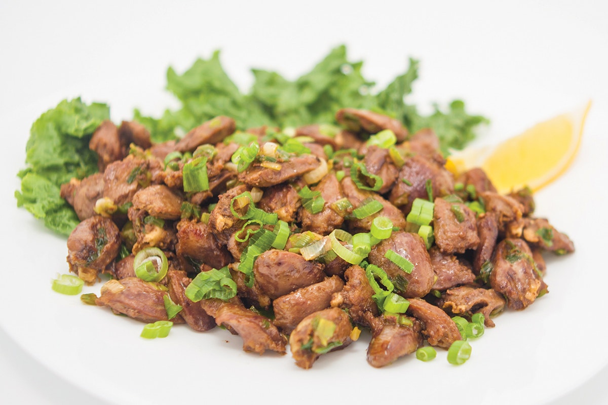 Generous serving of sauteed chicken hearts sitting on a white plate. Topped with sliced green onions and garnished with lemon wedge and curly leaf lettuce.