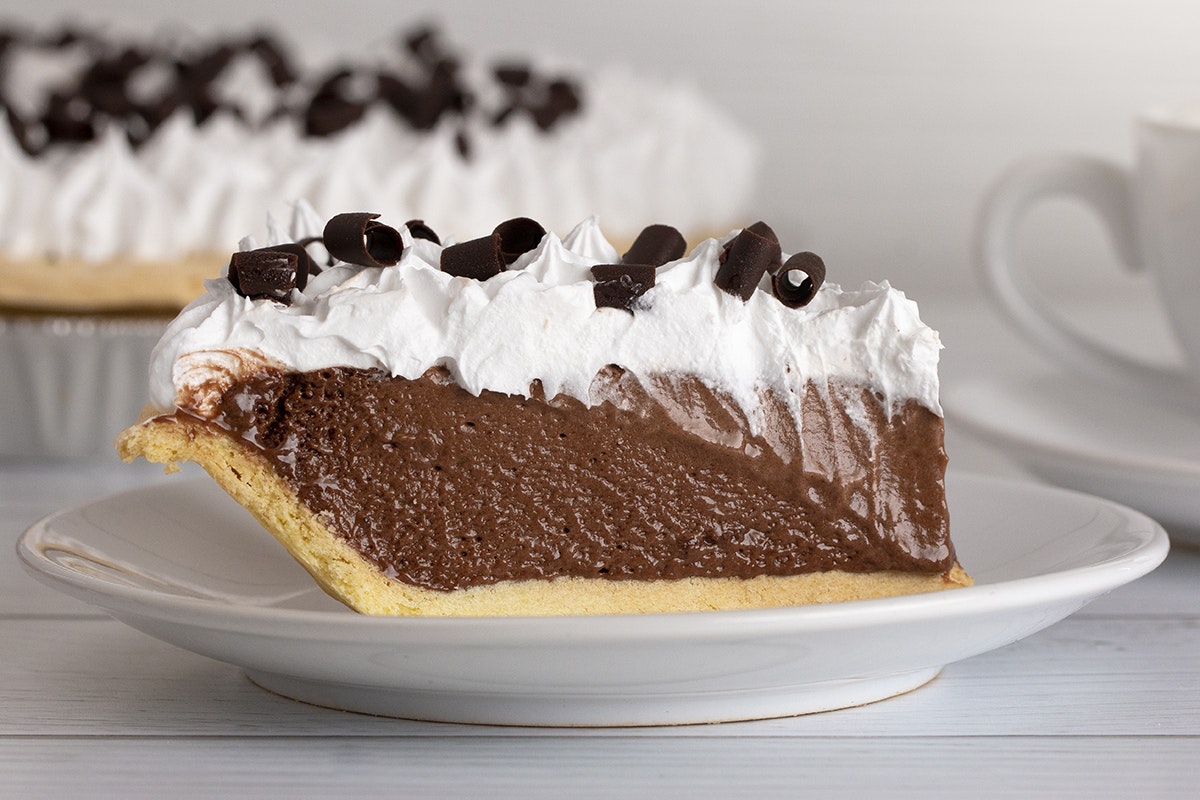 Slice of chocolate pudding pie, topped with whipped cream and chocolate shavings. On white plate, on white wood-grain table.