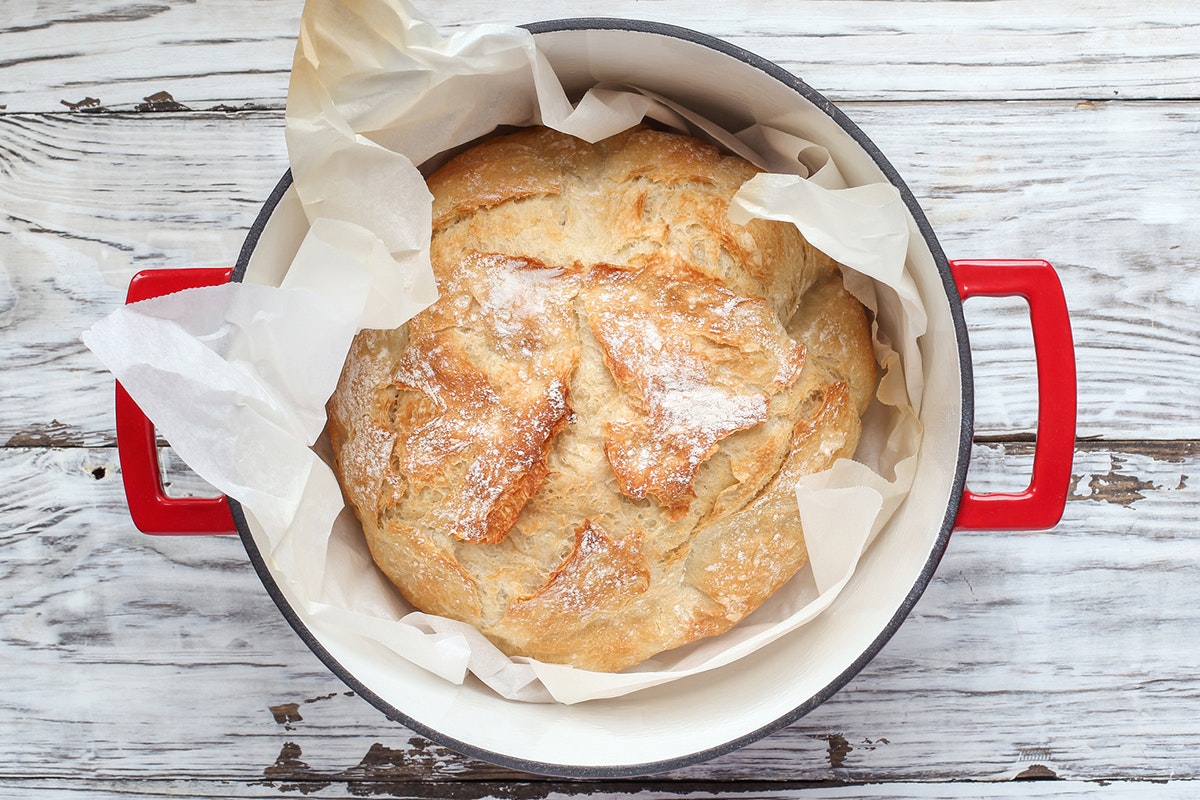 Round bread loaf sitting in a red Dutch oven pan. Parchment paper between bread and pan. White-painted wood grain surface behind.