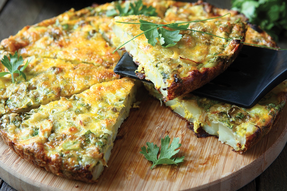 Round egg frittata filled with herbs and greens, cut into slices. Black spatula holding one slice above the rest, all on a circular wooden cutting board.