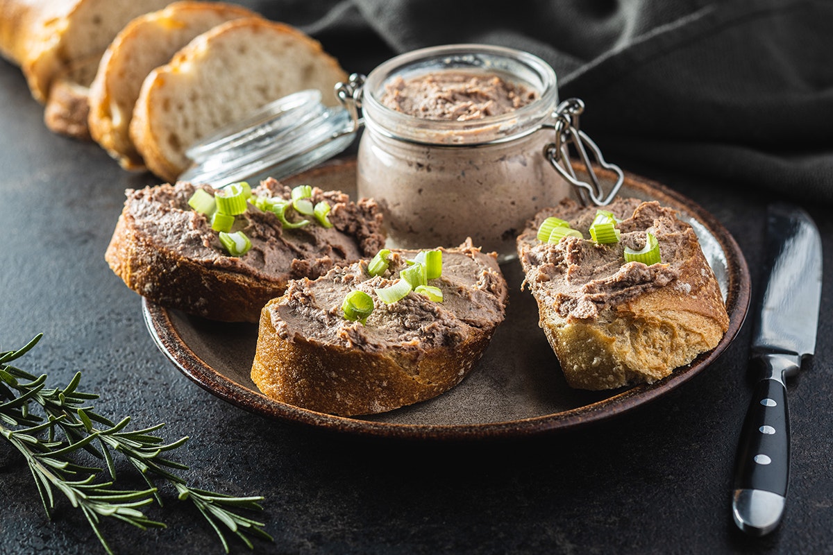 Liver pate spread on slices of french bread, topped with sliced green onions. Jar of pate alongside. Brown plate and fancy knife resting on dark gray surface.