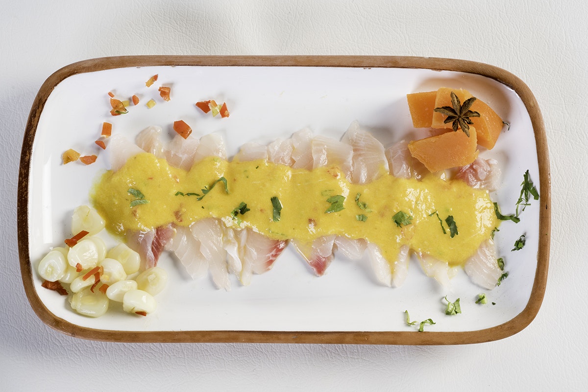 White and pink raw fish on a long platter, topped with light yellow sauce and accompanied by large corn kernels, orange sweet potato and other garnishes. White countertop.