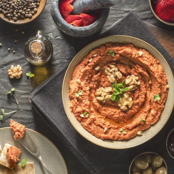 Dip made of red peppers and walnuts. Topped with walnut halves and herbs. Accompanied by bowls of olives and a plate with pita bread.