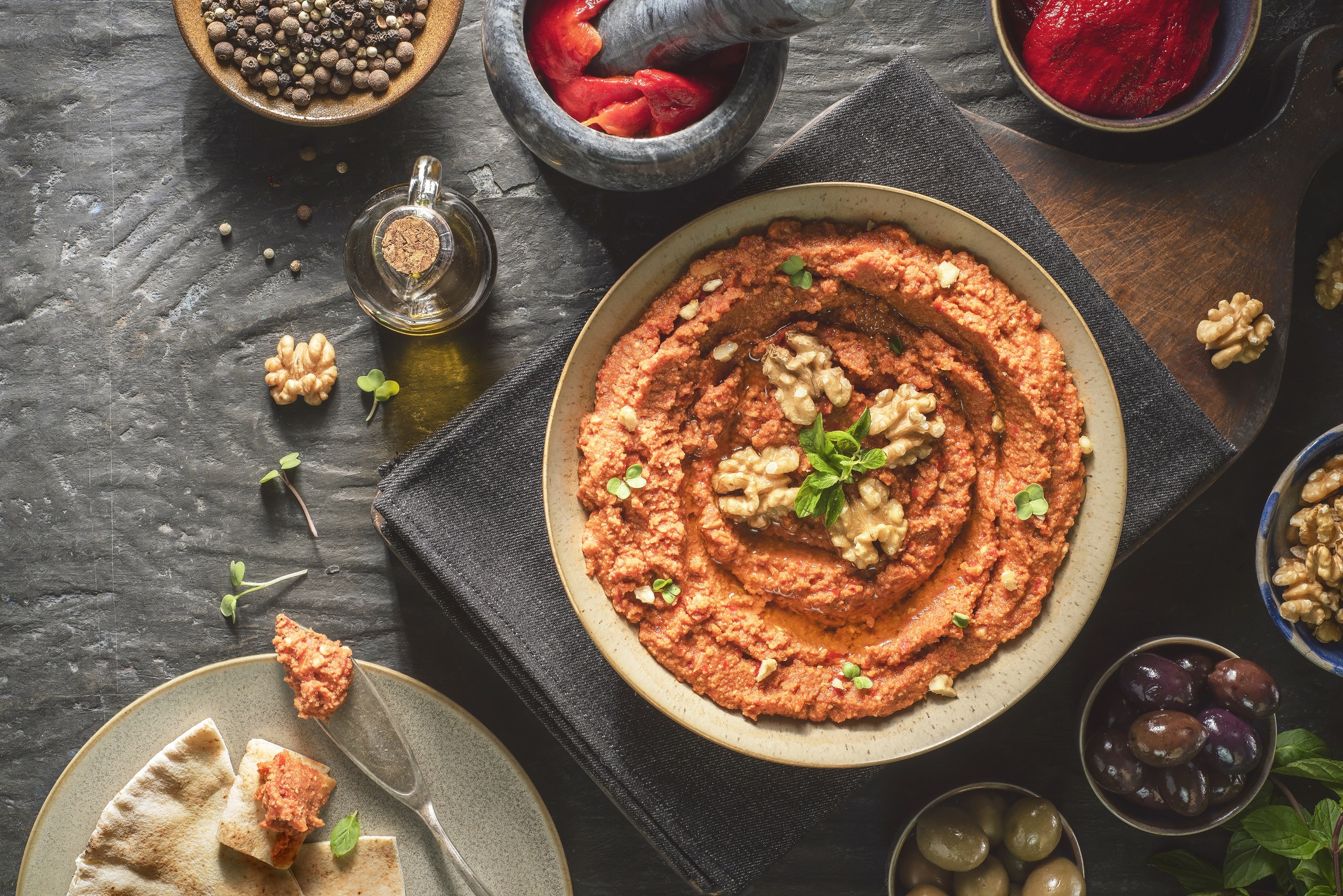 Dip made of red peppers and walnuts. Topped with walnut halves and herbs. Accompanied by bowls of olives and a plate with pita bread.