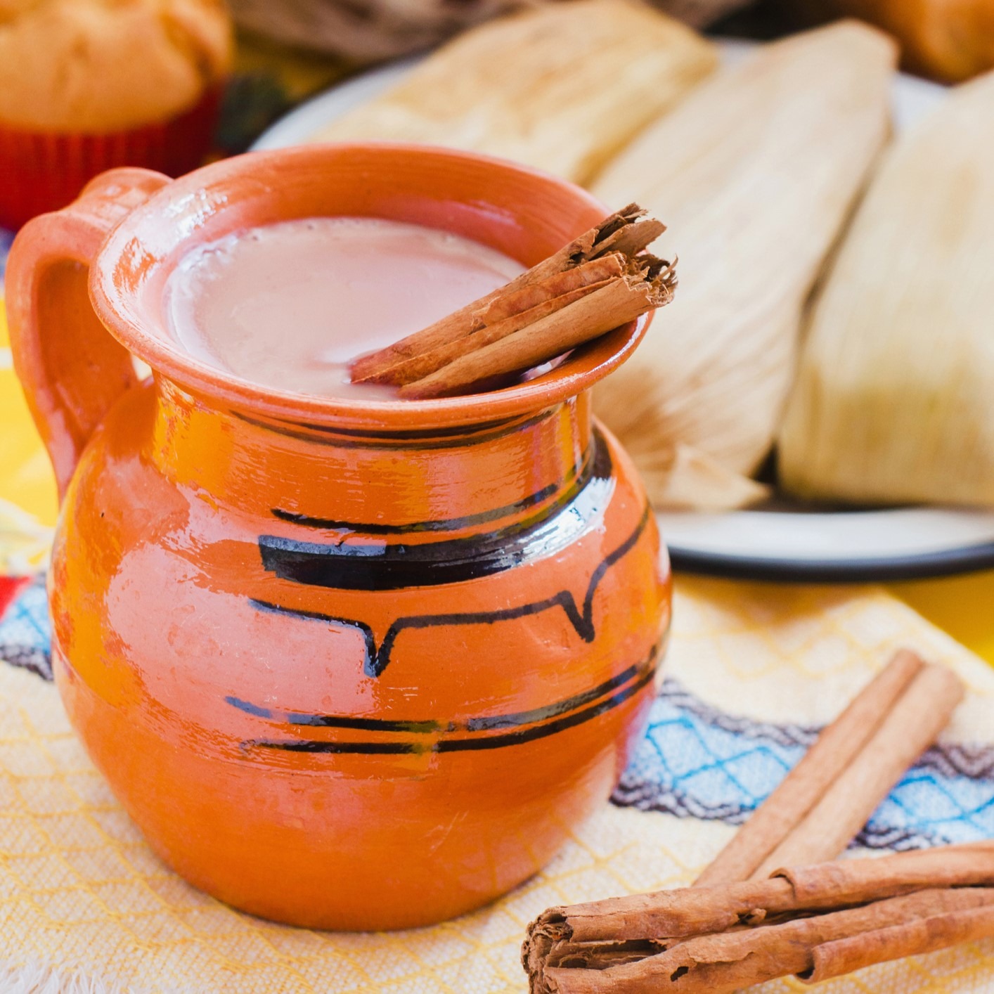 Mexican Atole - orange ceramic mug with cinnamon-cocoa drink. Cinnamon stick garnish and tamales in background.