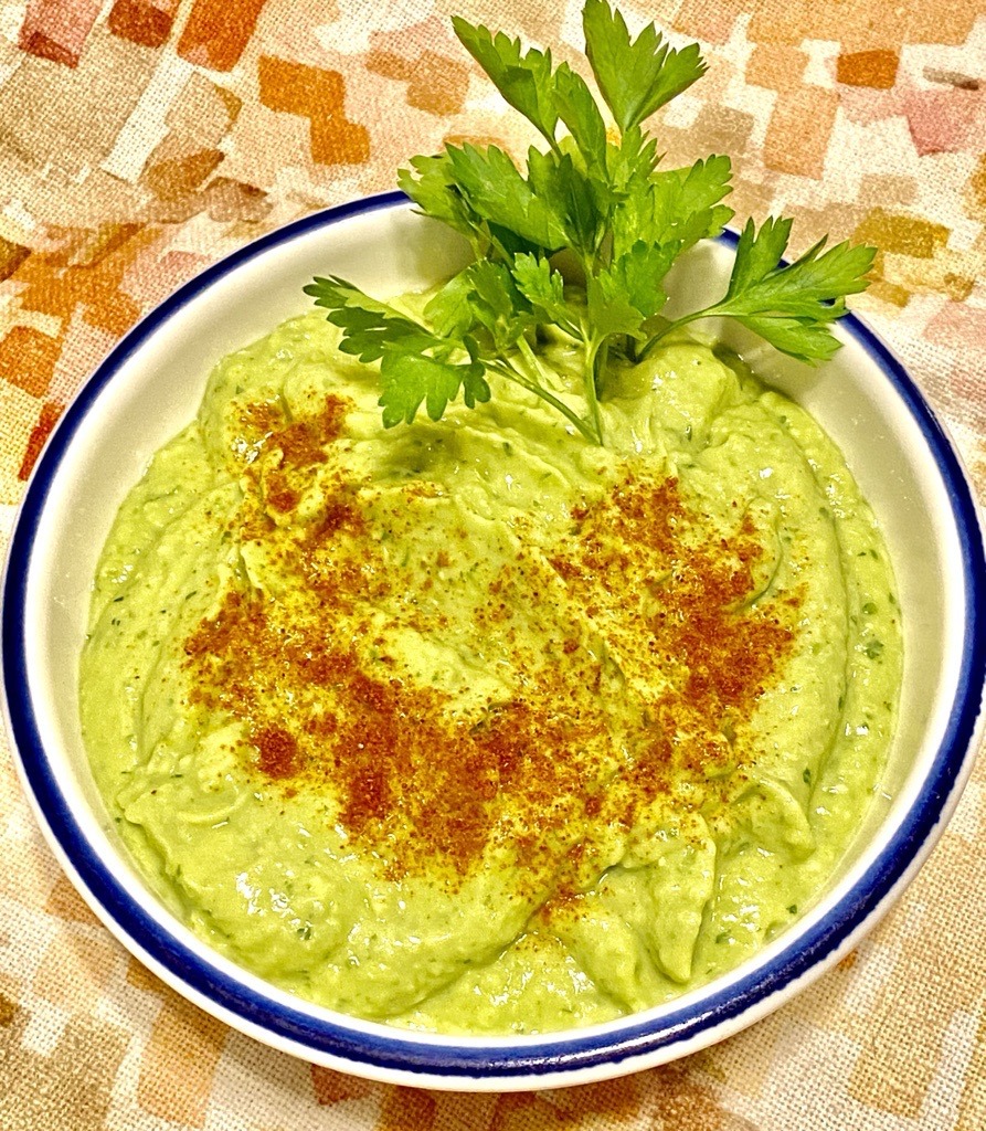 Green dip with darker green herb flecks, topped with cayenne pepper and garnished with parsley sprigs. White bowl with blue stripe. Multi-colored tablecloth.
