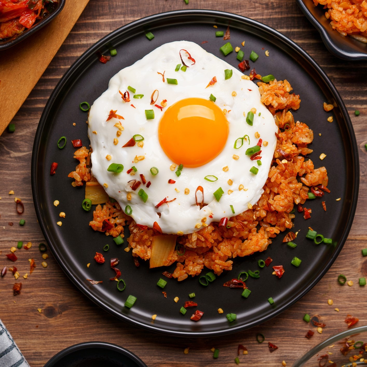 Fried rice with kimchi. Topped with fried egg, scallions and chili flakes. Black plate, dark wood background.