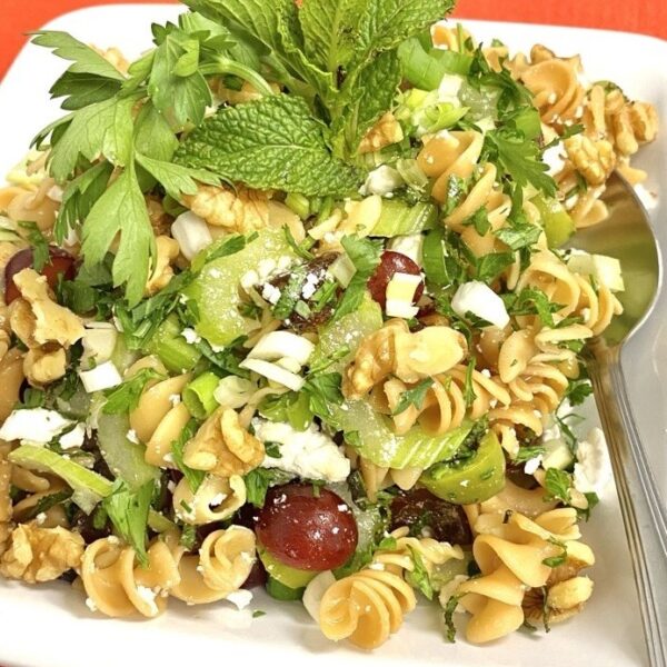 Pasta Salad with Celery, Dates, Feta and Fresh Herbs. Also red grapes and green olives, garnished with mint/parsley. Spoon resting to the side. Orange background.