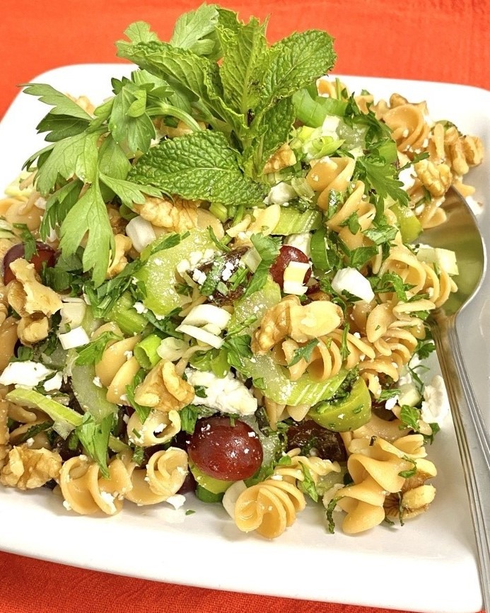 Pasta Salad with Celery, Dates, Feta and Fresh Herbs. Also red grapes and green olives, garnished with mint/parsley. Spoon resting to the side. Orange background.