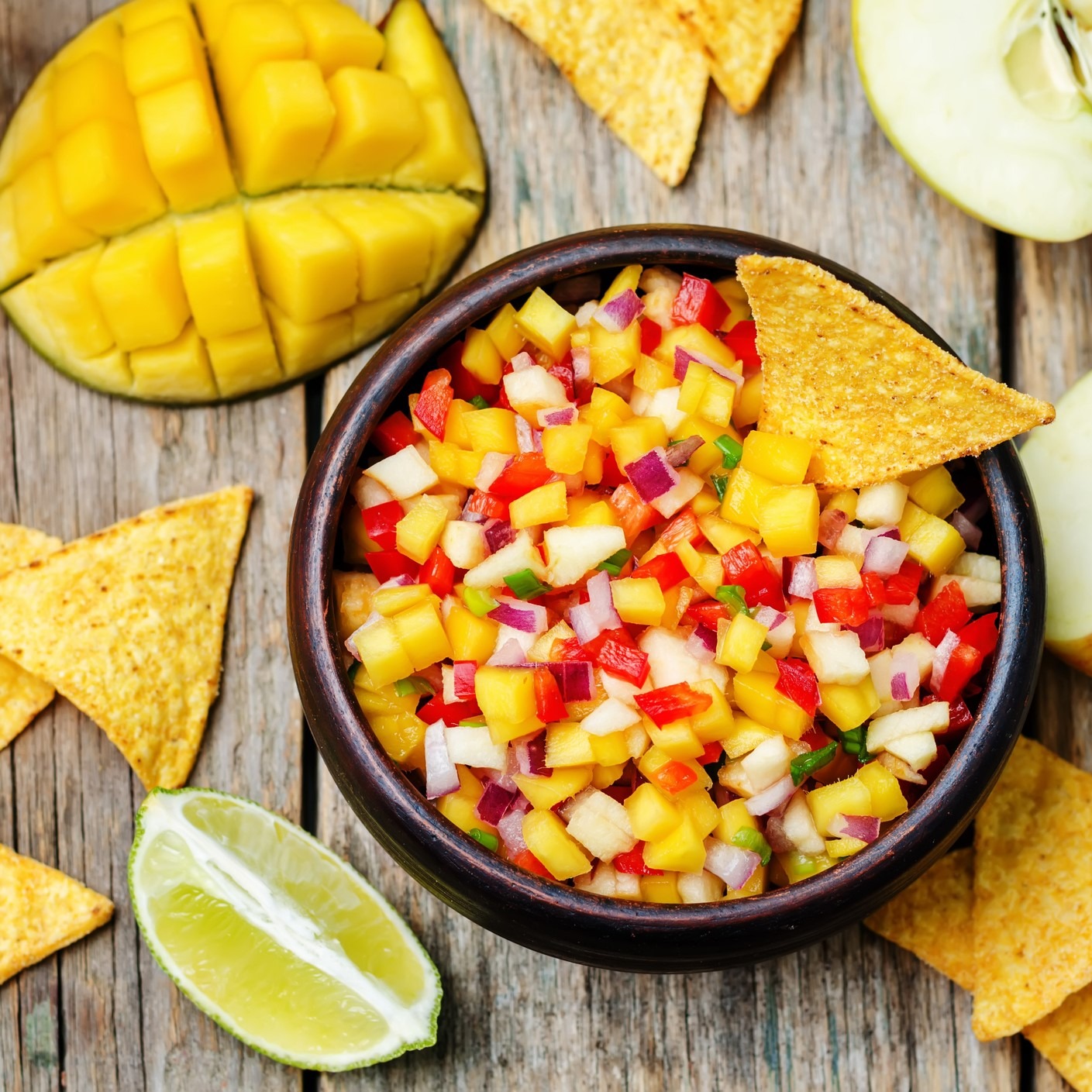 Salsa of apples, mango, onion, cliantro, jalapenos. Round brown bowl surrounded by corn chips, lime/mango/apple slices. Wooden table.