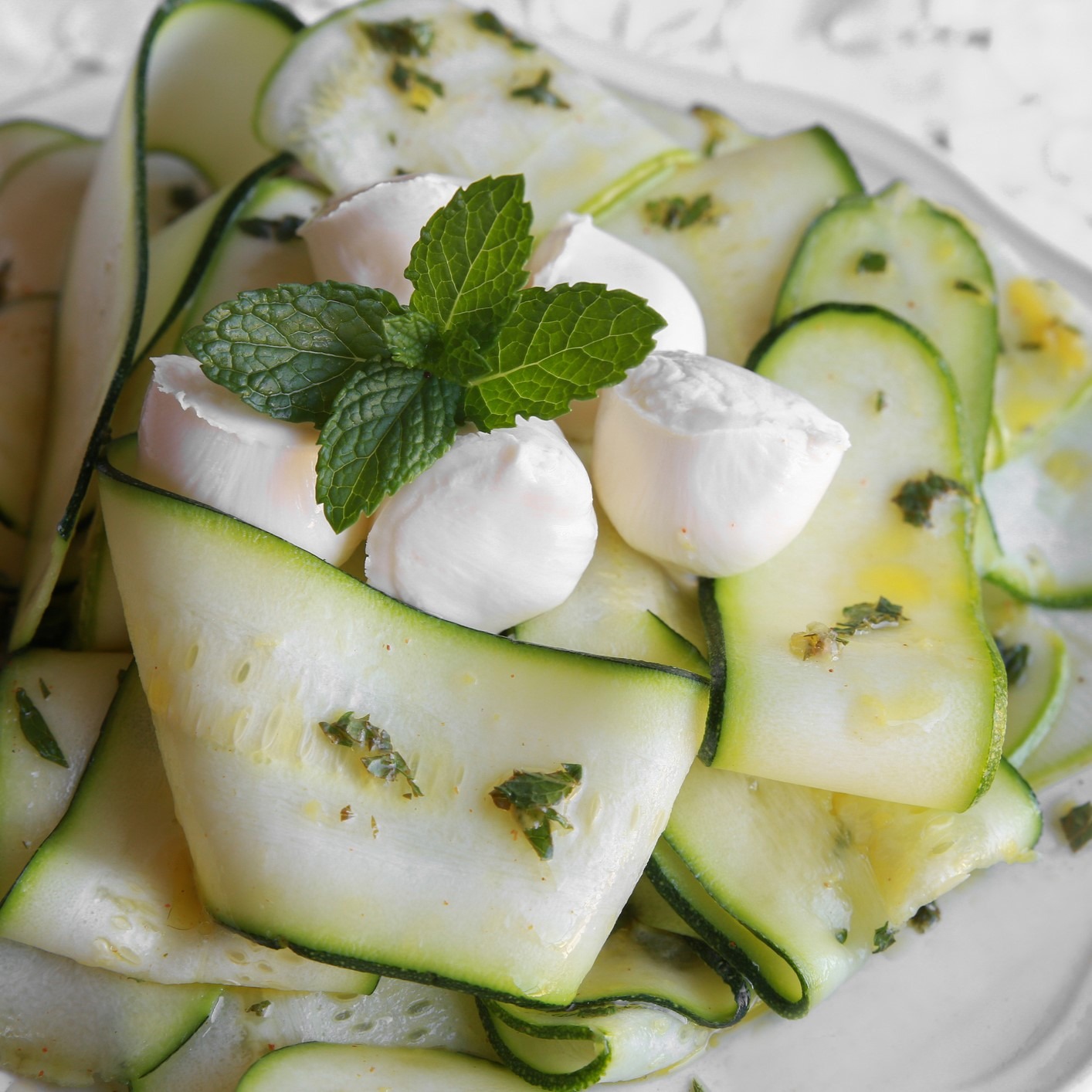 Layers of thinly-sliced zucchini dressed with olive oil and topped with mint and fresh mozzarella. White plate.