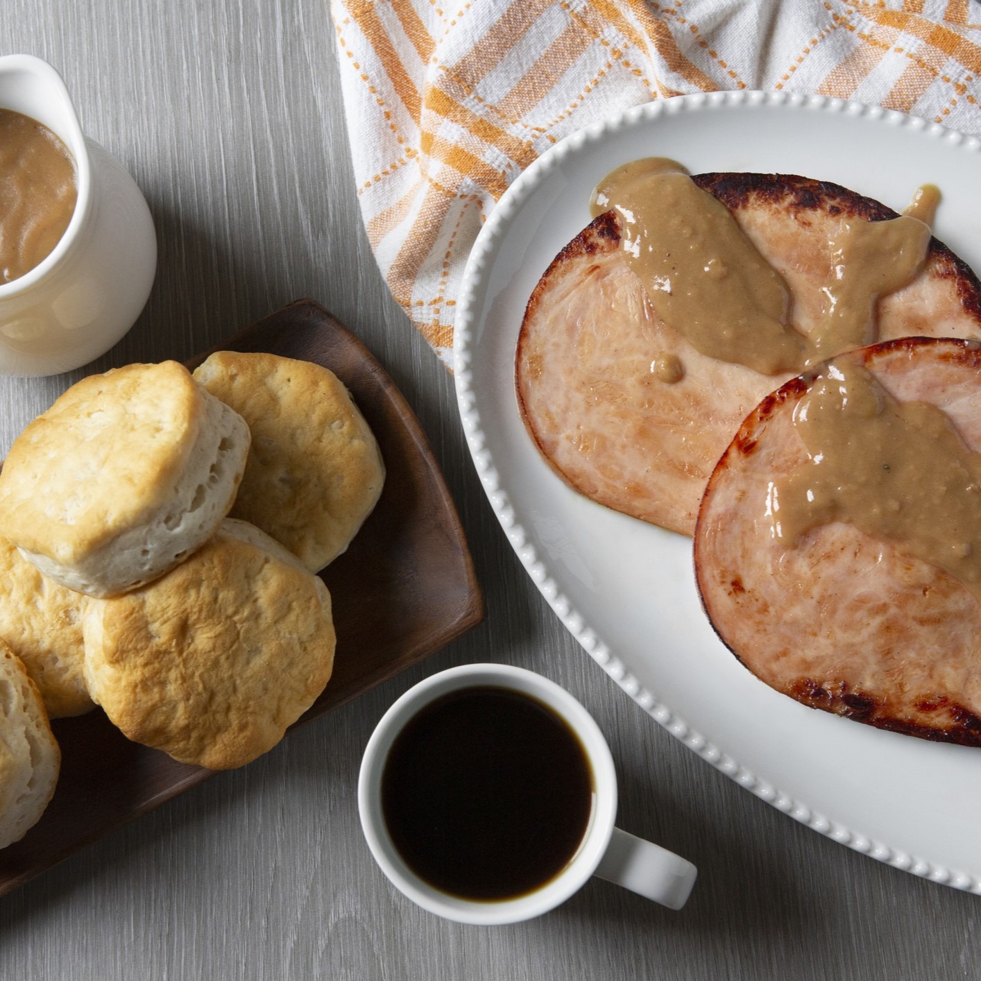 White platter with ham steaks drizzled with gravy, next to another platter with biscuits. Cup of coffee and a small gravy pitcher on the side. Gray woodgrain table and white napkin with maize yellow plaid detail.