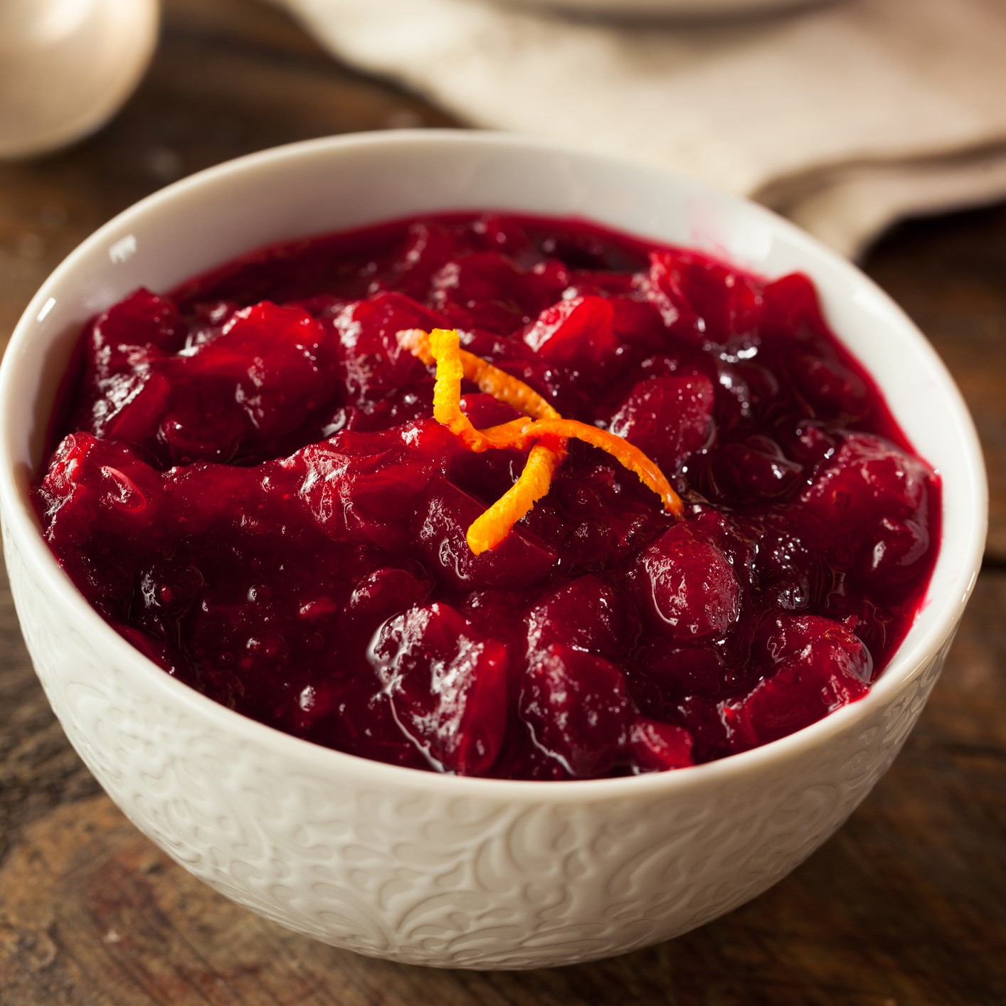 White bowl with cranberry relish. Garnished with orange zest. Atop a brown wooden table.