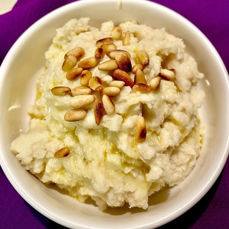 Mashed potatoes topped with olive oil and toasted pine nuts. White bowl and purple tablecloth.