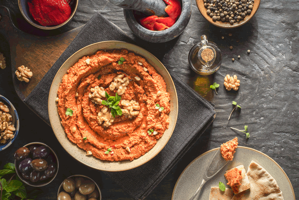 Muhammara middle eastern red pepper dip with walnuts and fresh herbs on a black slate background with red peppers, green and black olives and black peppercorns.