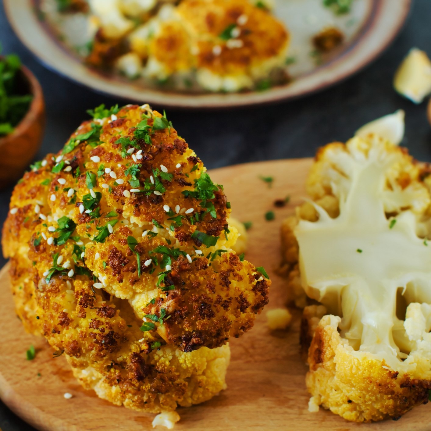 Whole roasted head of cauliflower with sesam seeds and parsley garnish. On round wooden cutting board.