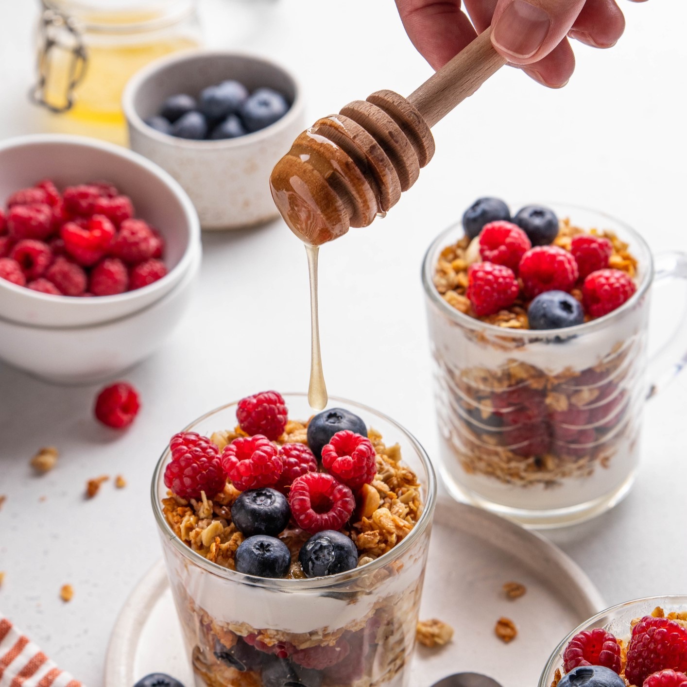 Layers of white yogurt, granola and fresh berries in clear glass mugs. Drizzle of honey coming from above. Bowls of additional berries in background.