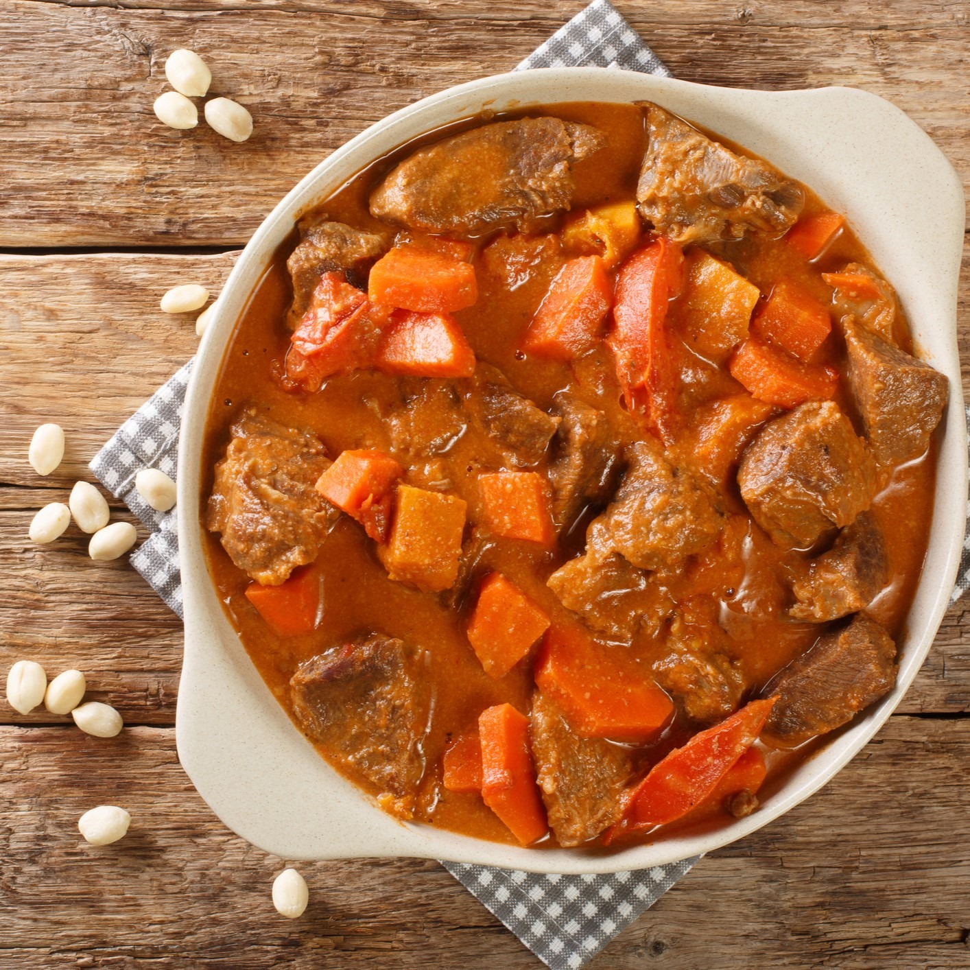 Beef stew with vegetables in white casserole dish sitting on white/gray checked napkin and wooden table.