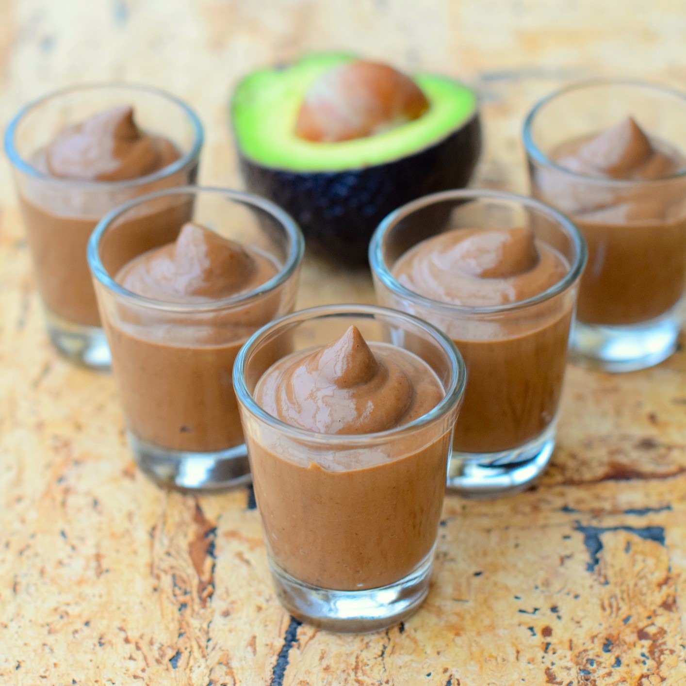 Chocolate mousse in small glasses, on wooden surface with avocado half in background.