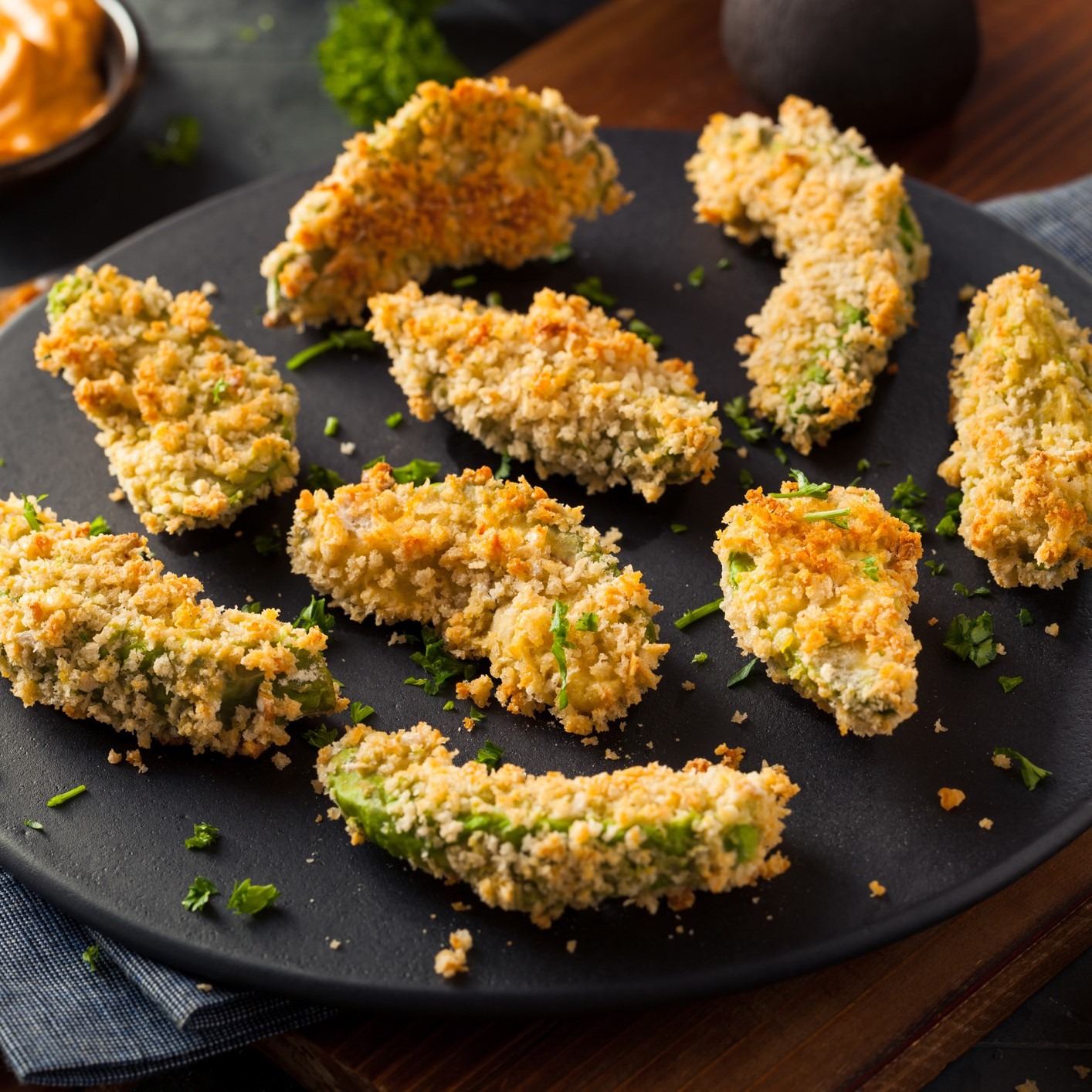 Avocado wedges covered in breadcrumbs and oven-baked. Topped with fresh chopped herbs on black serving plate.