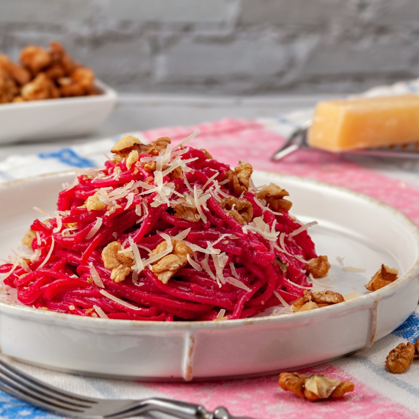 Beet Pasta - noodles with bright pink sauce, topped with shredded cheese and toasted walnuts. White bowl, colorful tablecloth.