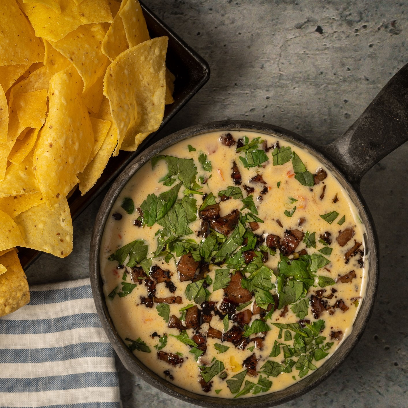 Serving dish of queso dip with meat and cooked onions. Topped with chopped fresh herbs, served with corn chips. Black serving bowl with handle.