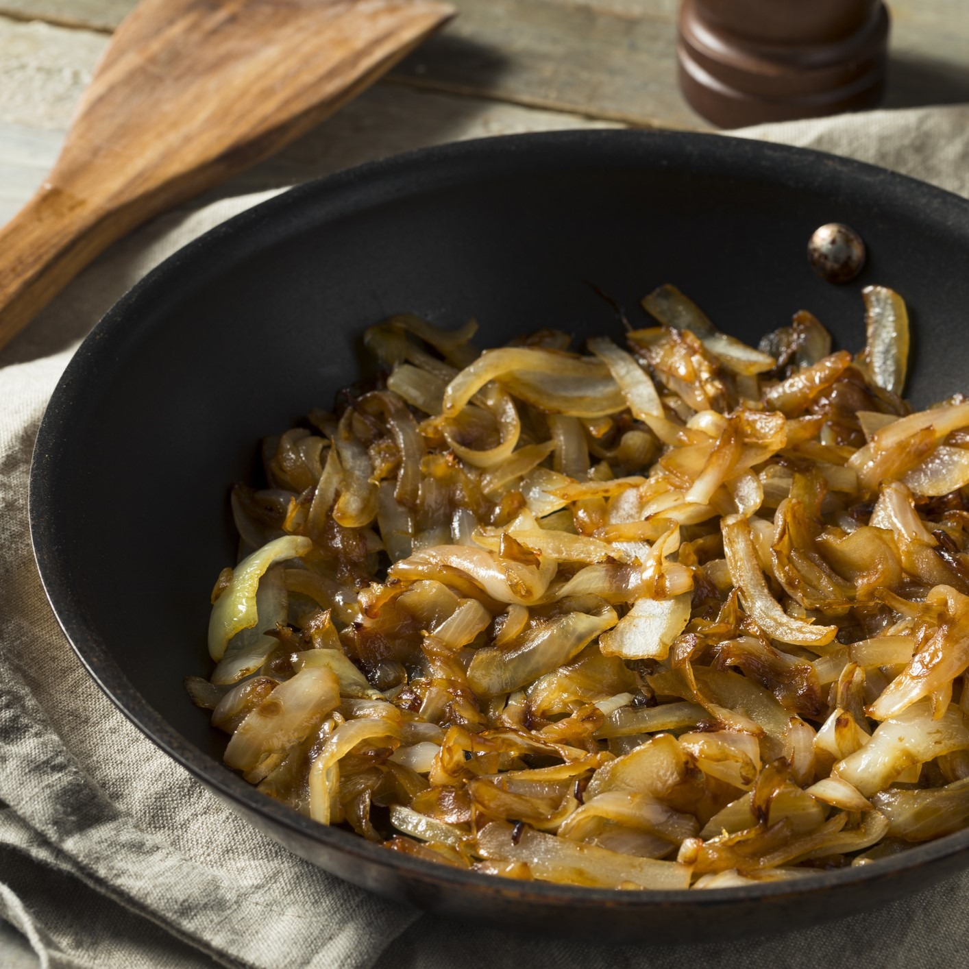 Saute pan with caramelized onions. Sitting on tan tablecloth, wooden spatula nearby.