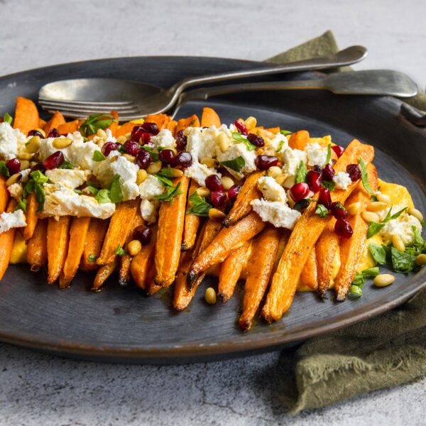 Roasted root vegetables lined up and topped with goat cheese, pomegranate arils and fresh herbs. Gray ceramic plate, metal serving utensils and dark green napkin.