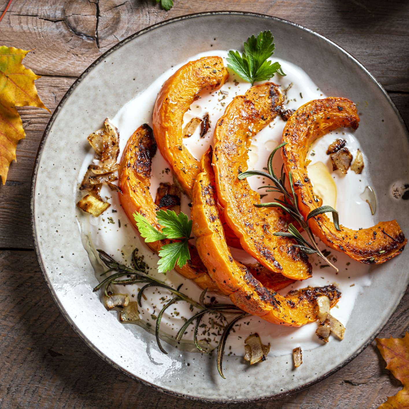 Roasted kabocha squash with sweet-spicy glaze, roasted herbs and leeks. On a bed of Greek yogurt sauce and garnished with fresh parsley. Round, gray ceramic plate on wooden surface.