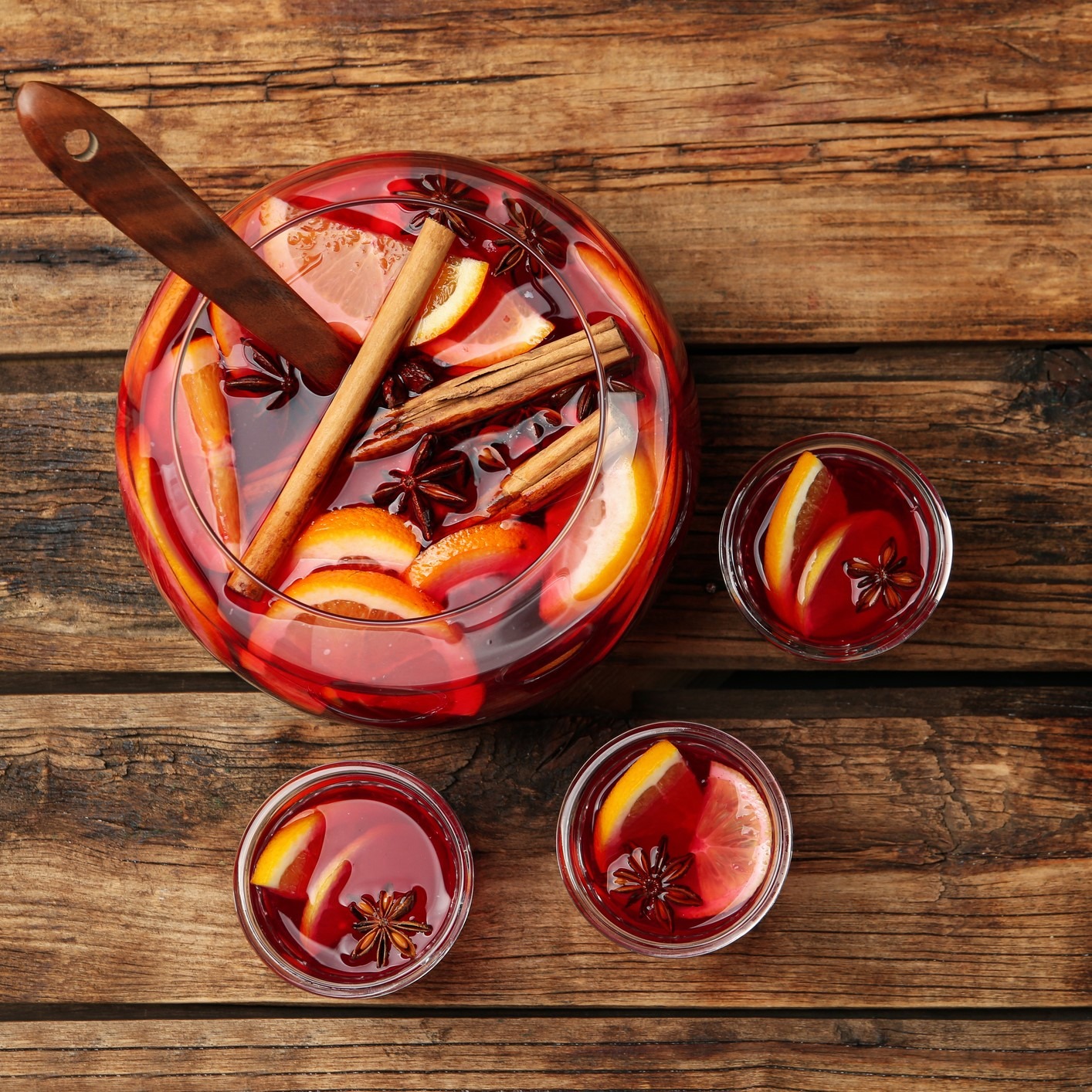 Mulled wine with whole spices and orange slices. Serving bowl with wooden spoon, sitting next to individual glasses on a wooden table.