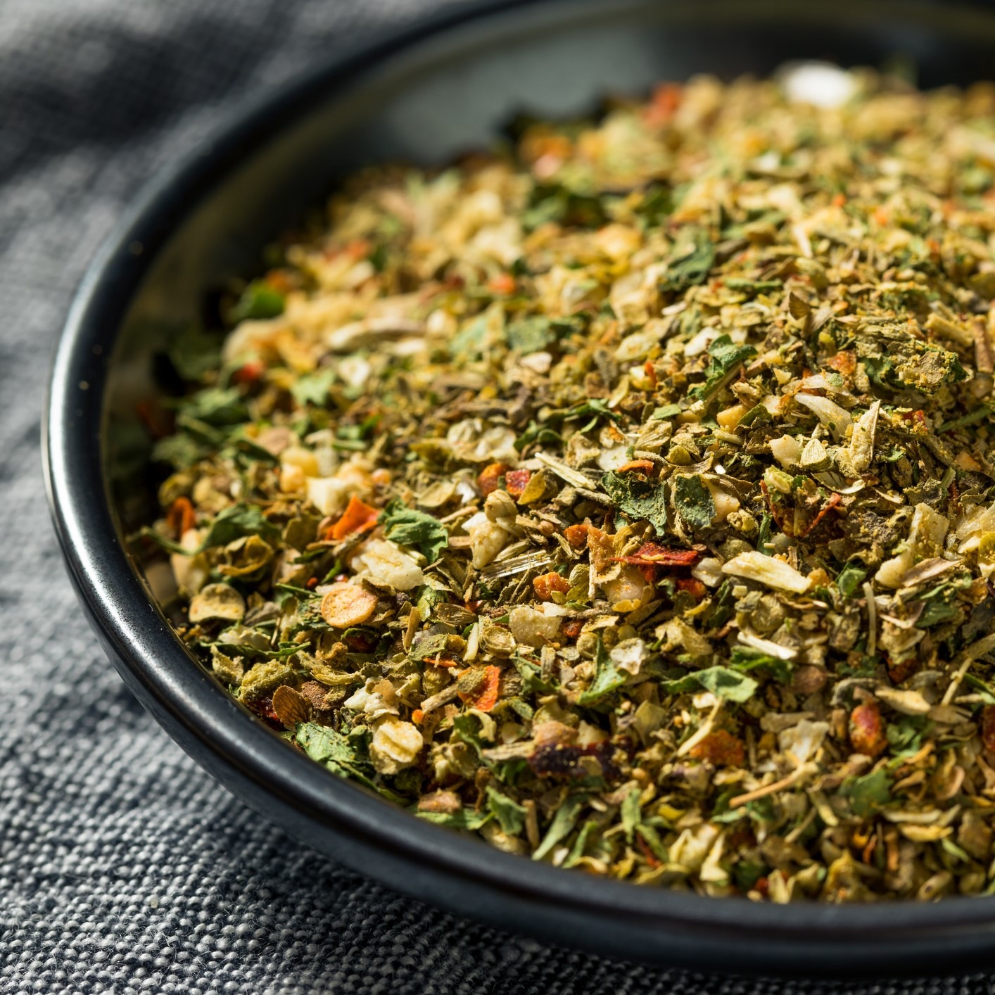 Combination of dried herbs and ground spices in black bowl on gray tablecloth.