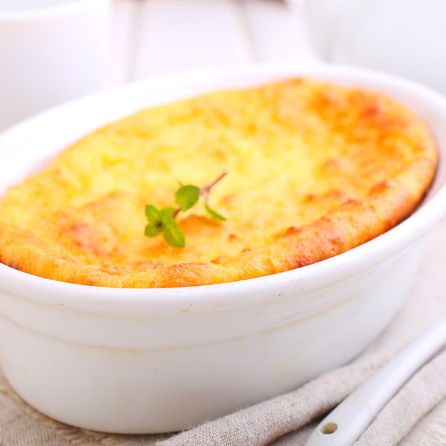 Corn spoonbread in an oval-shaped white casserole dish. Tan placemat and wooden serving spoon.