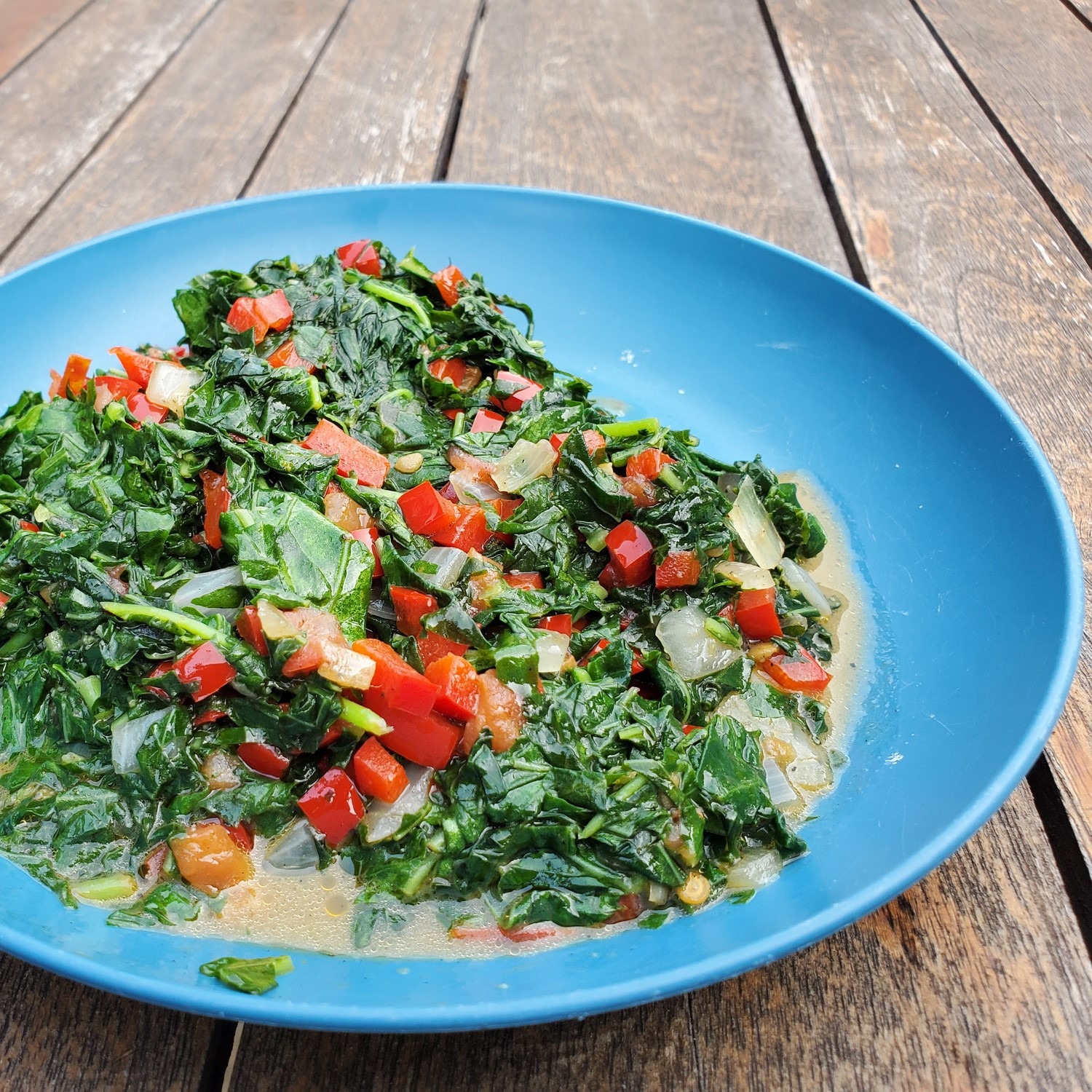 Braised greens with onions and tomatoes in a bright blue serving bowl. Wooden table.