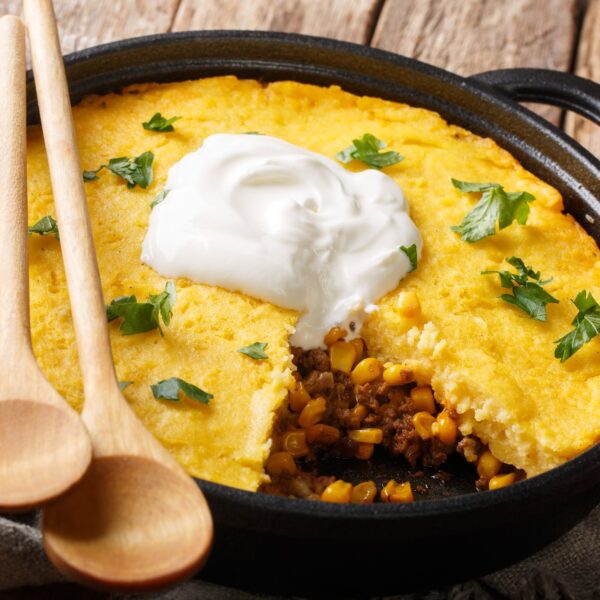 Tamale Pie Casserole - beef and corn - in iron pan. Topped with sour cream and fresh herbs. Wooden serving spoons perched on side of pan.