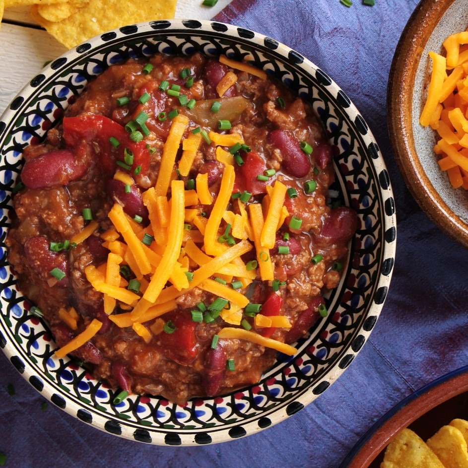 Ground meat chili with kidney beans and vegetables. Topped with shredded cheddar cheese and chopped chives. Ceramic bowl with colorful design. Blue tablecloth.