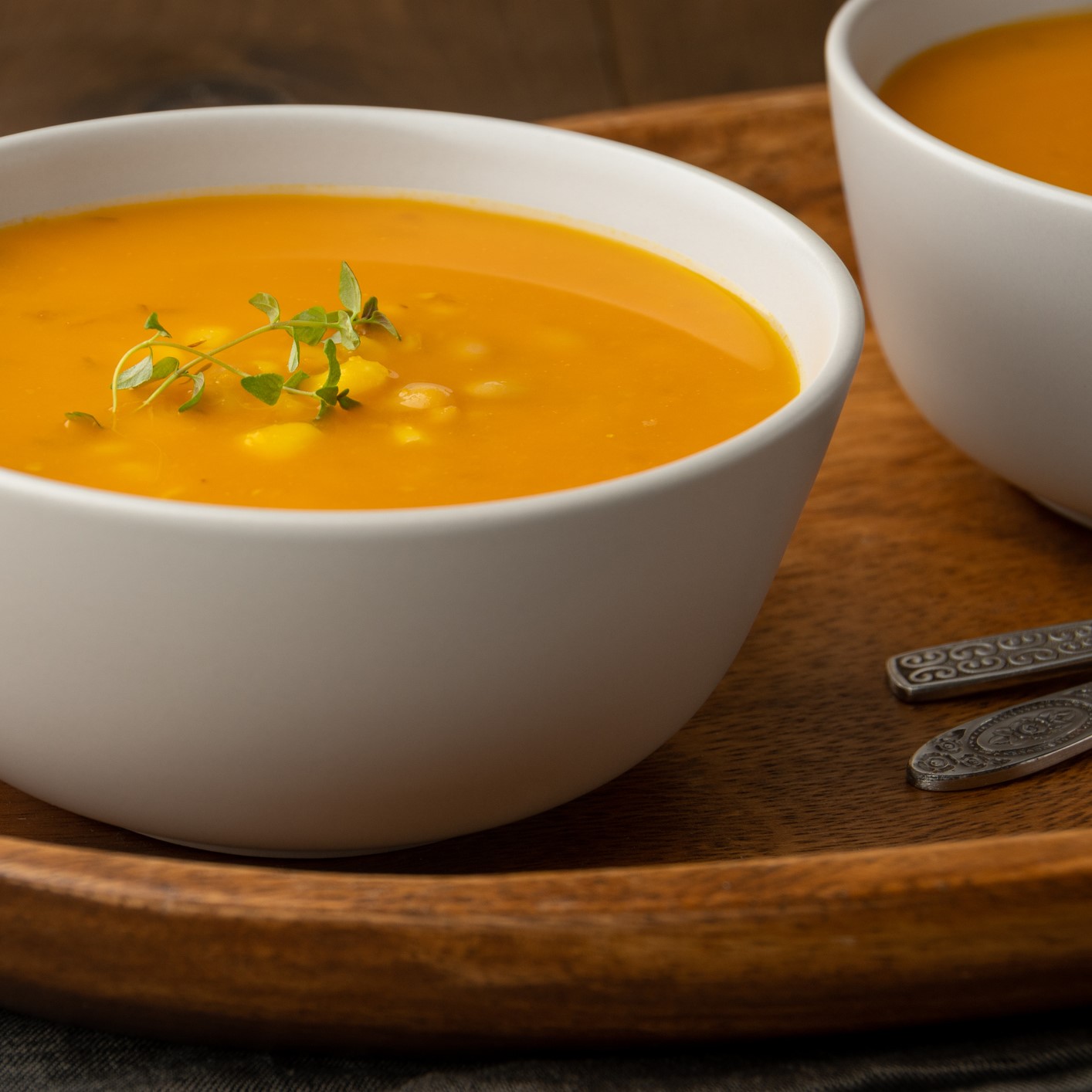 Whie ceramic bowl of orange squash soup with hominy and fresh herbs. Wooden tray.