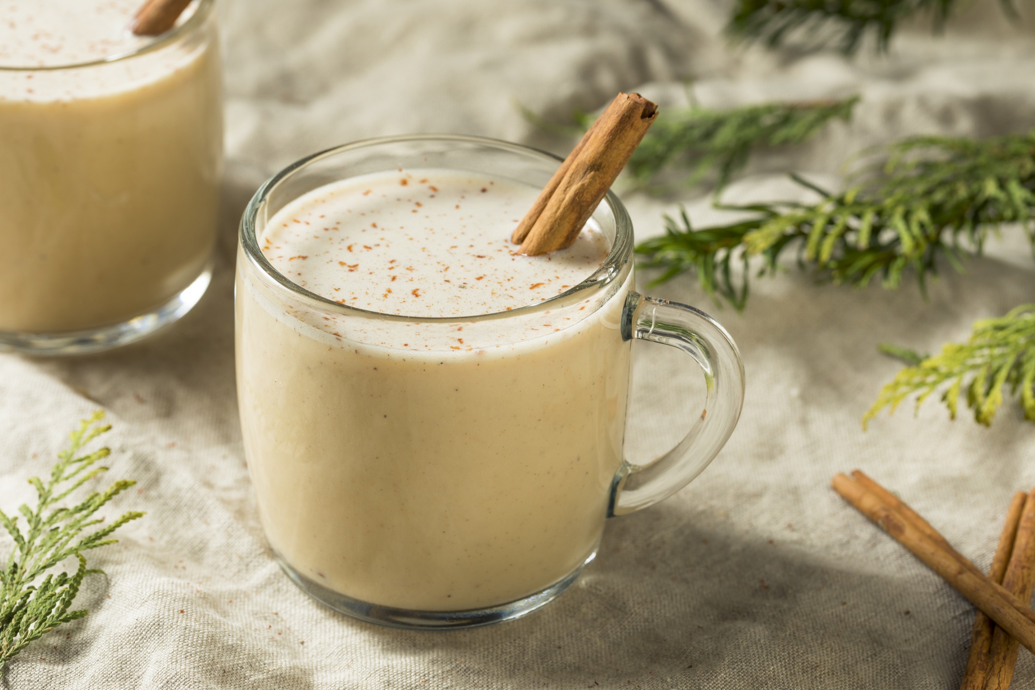 Clear glass mug of Coquito, accented with cinnamon stick and flecks of ground cinnamon. Pine branches for holiday cheer.