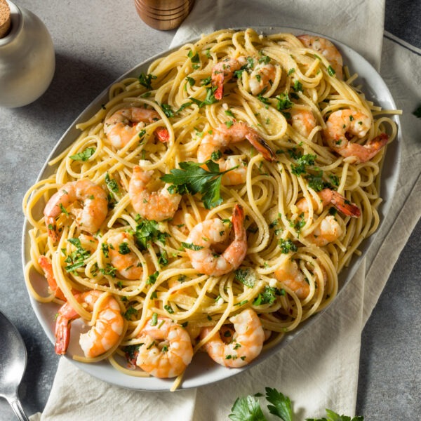 Shrimp Scampi with pasta and fresh herbs in a large oval serving bowl. Gray tabletop with linen napkin and serving spoon.