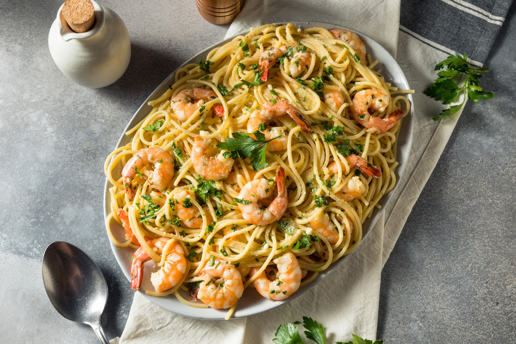 Shrimp Scampi with pasta and fresh herbs in a large oval serving bowl. Gray tabletop with linen napkin and serving spoon.