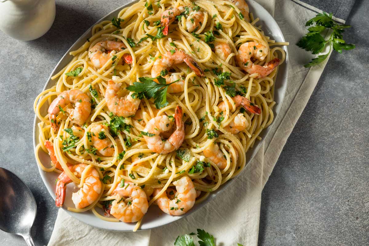 Shrimp Scampi with pasta and fresh herbs in a large oval serving bowl. Gray tabletop with linen napkin and serving spoon.