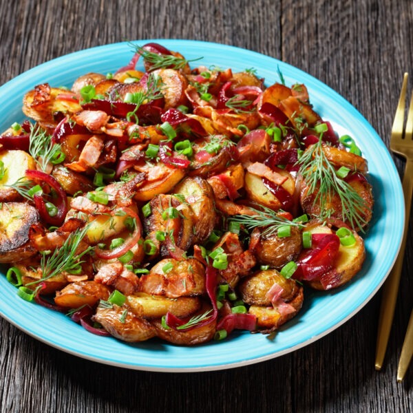 Salad of roast potato salad with red onion, bacon and fresh chopped herbs on a blue plate on a dark rustic wooden table.